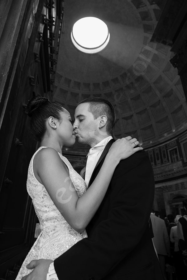 Kissing at the Roman Pantheon by the entrance of the structure. Light shining through the hole. 