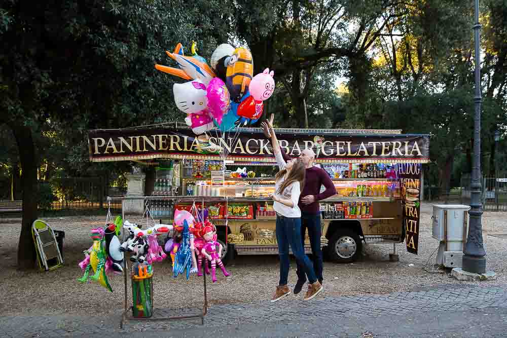 Couple jumping up in the air trying to catch some balloons in front of a gelato stand.