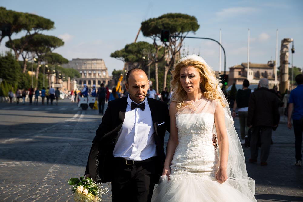 Photo reportage style photography as newlyweds walk on via dei Fori Imperiali with the Colosseum in the background. Post wedding photography.