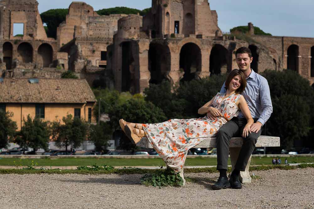 Portrait image of a couple sitting down with the Palatine hill in the background. Rome Engagement couple. 