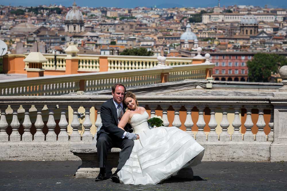 Newlywed couple sitting down on a marble bench before the roman rooftops