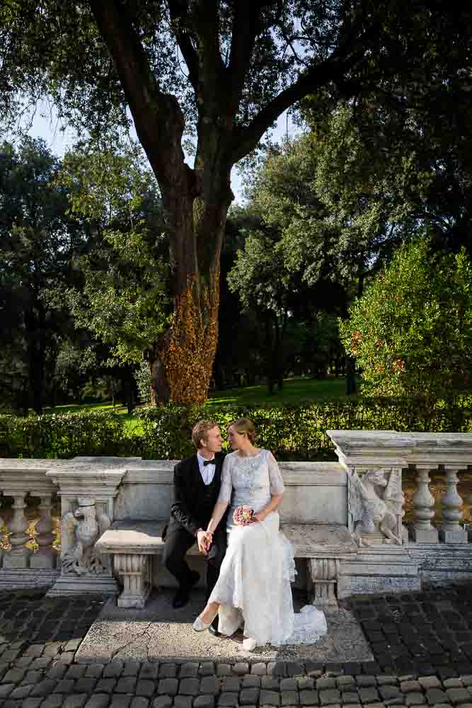 Portrait picture of a married couple sitting on a marble bench. Under Villa Borghese park trees.