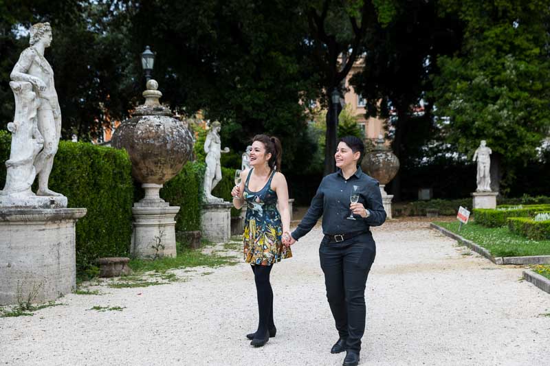 Same sex couple walking together in a roman park with ancient marble statues on the sides