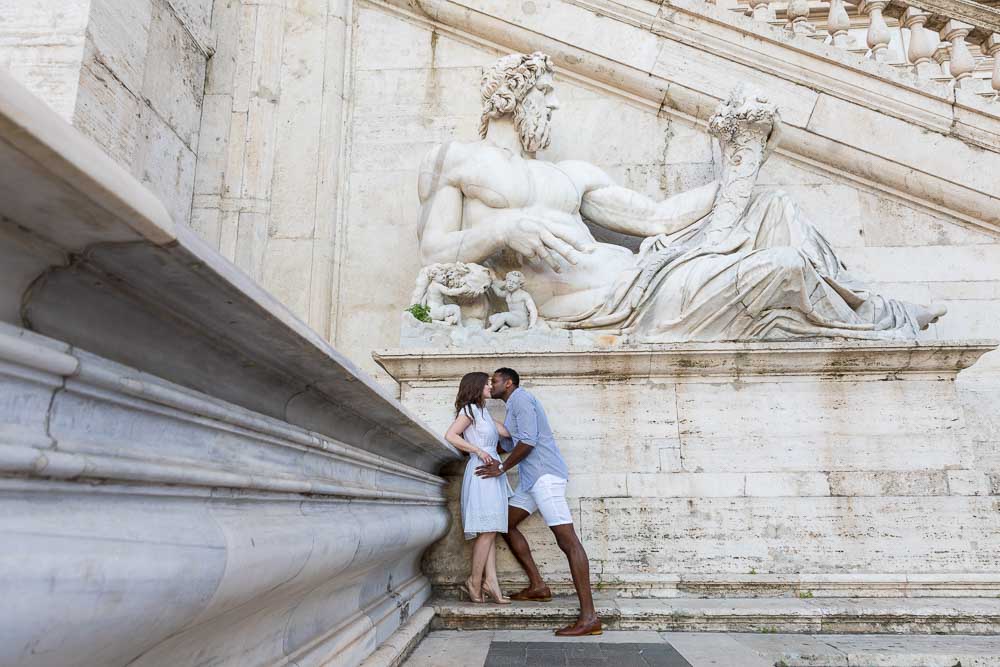 Portrait picture kissing under marble roman statue in Piazza del Campidoglio