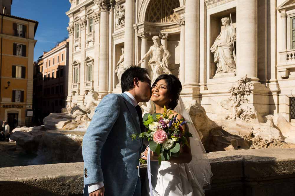 Groom kissing Bride in front of the Trevi fountain in soft midday sunlight