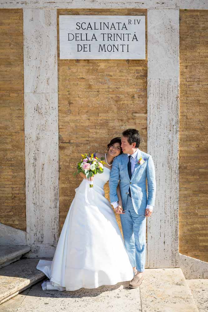 Newlyweds standing and posing together while leaning against the wall beneath the Trinita dei Monti sign