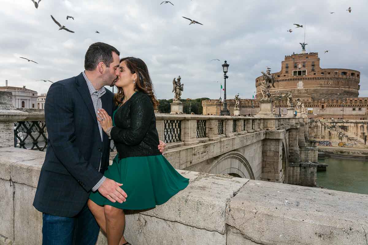 Kissing with Castel Sant'Angelo bridge in the distance and Castel St Angelo as backdrop with birds flying all around