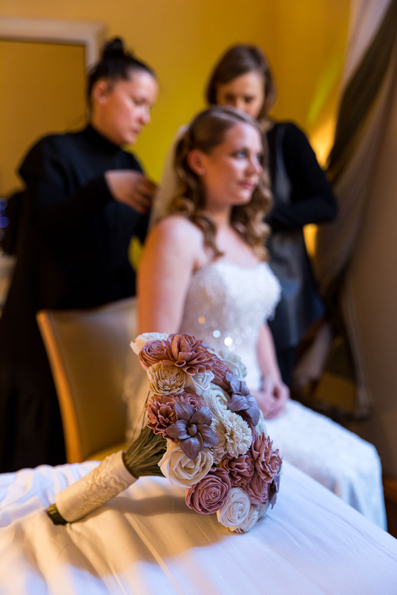 Bridal bouquet in the forefront in focus with the bride and the makeup team in the background out of focus