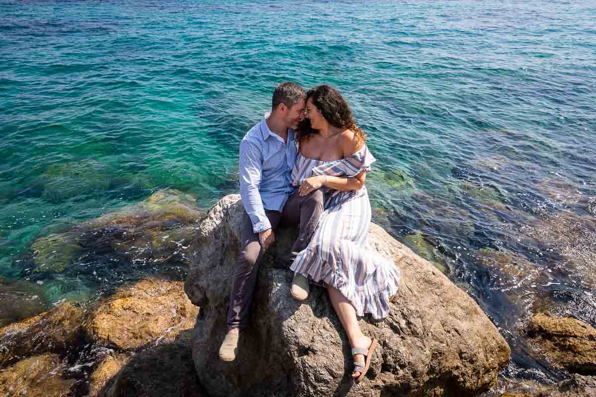 Capri engagement photo session. Couple sitting down on the rocks surrounded by crystalline emerald green blue water all around