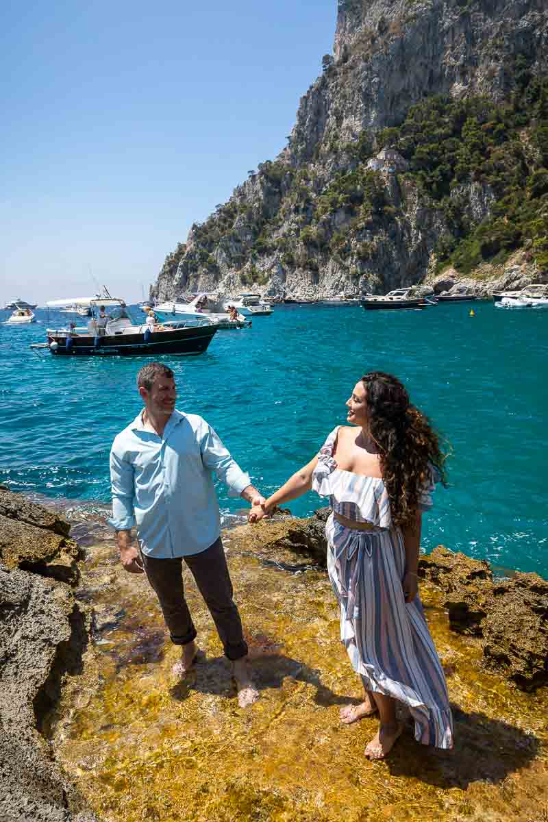 Couple holding hands and looking at each other while standing on yellow rocks with the beautiful Mediterranean sea as background. Capri Engagement Photo Session