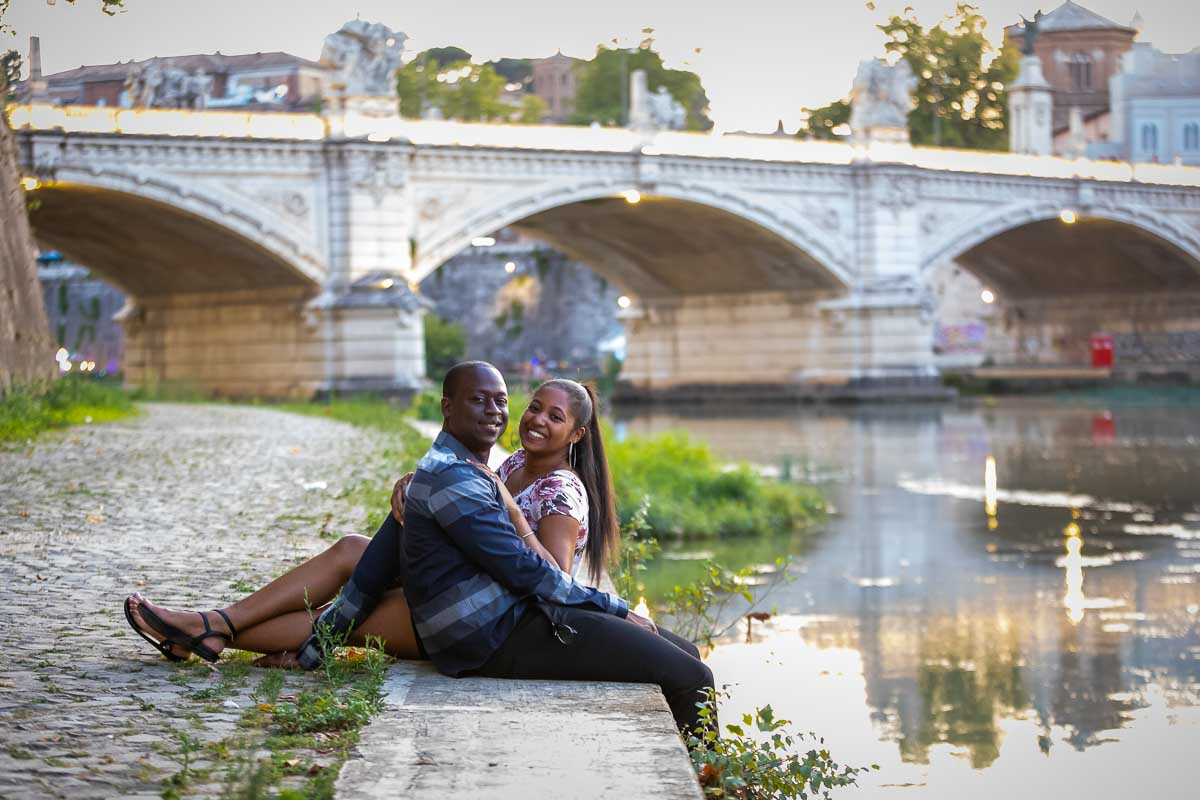 Couple posing while sitting down next to the river water with lit bridge behind them