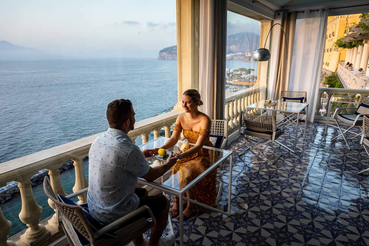 Holding hands while sitting at a table taking engagement pictures together and outside the beautiful seaside view