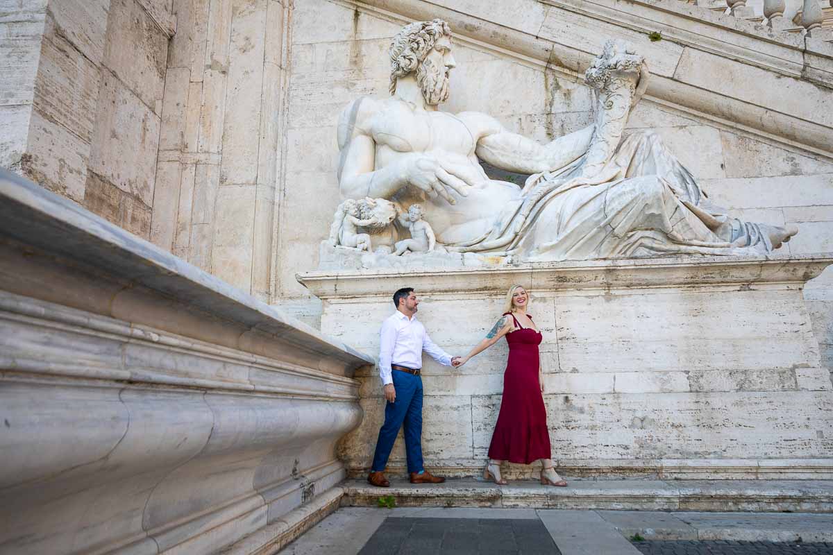 Walking picture of a couple leading while photographed underneath a large marble roman statue