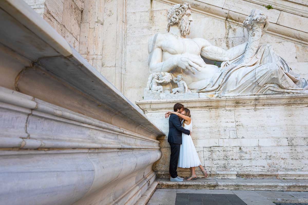 Posed image of groom and bride kissing underneath a large white marble roman statue 