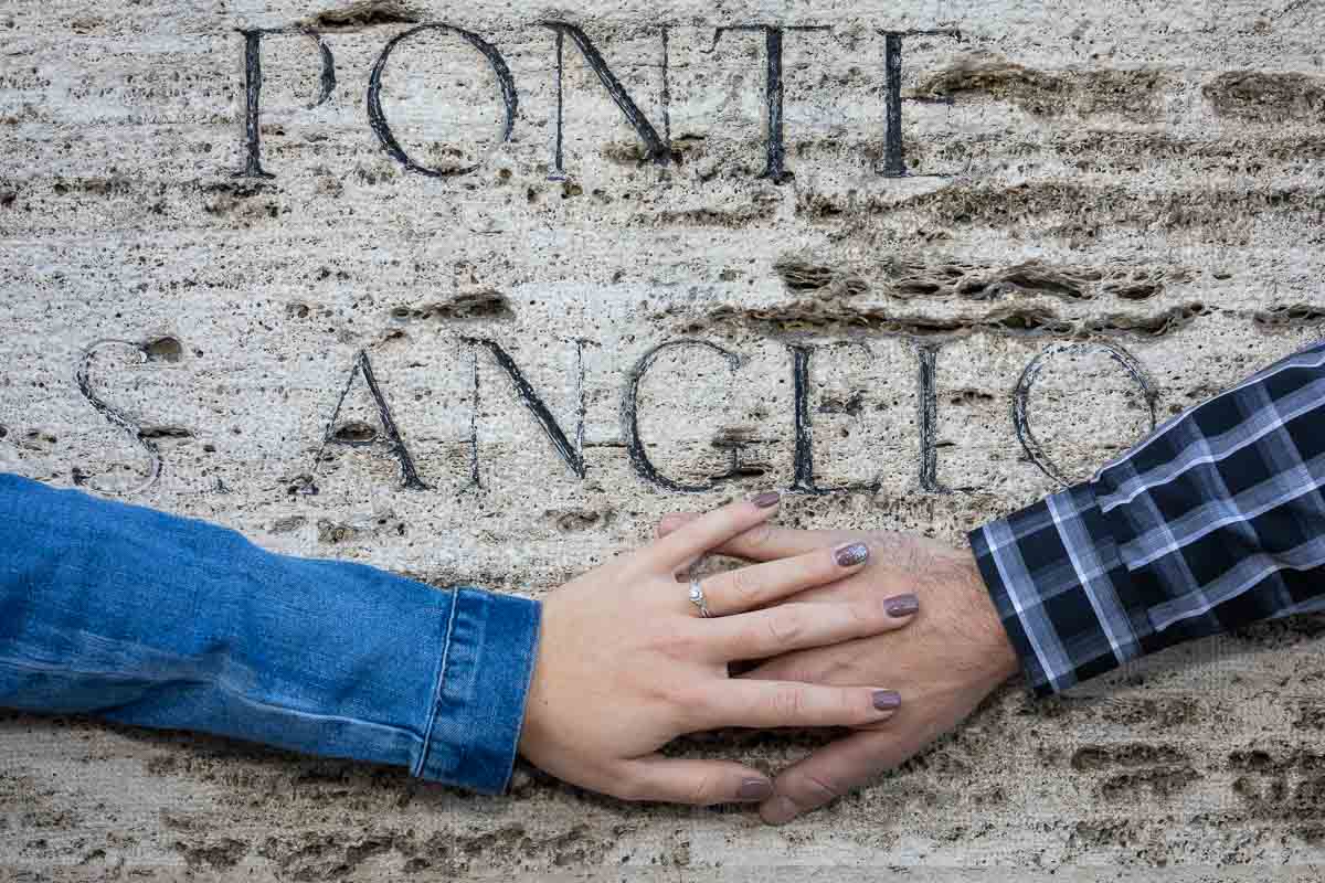 Photo of overlapping hands showing the beautiful engagement ring photographed against peperino rock with the name of ponte s. angelo