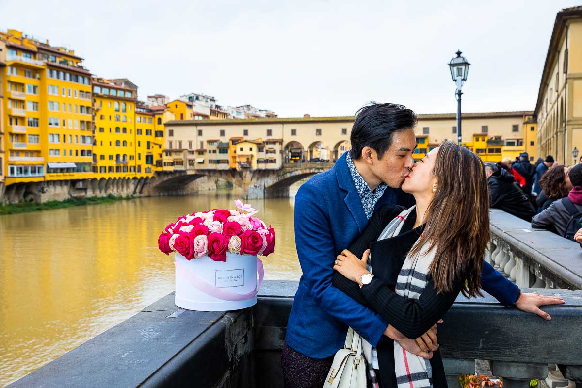 Couple kissing just engaged over the Arno river with Ponte Vecchio bridge in the far distance. In love in Florence Couple PhotoShoot