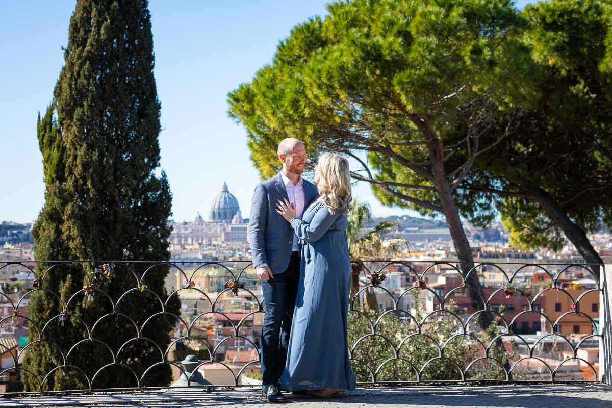 Couple standing together with Saint Peter's dome cathedral in the far distance in front of Mediterranean stone pine trees. Surprise Engagement Rome