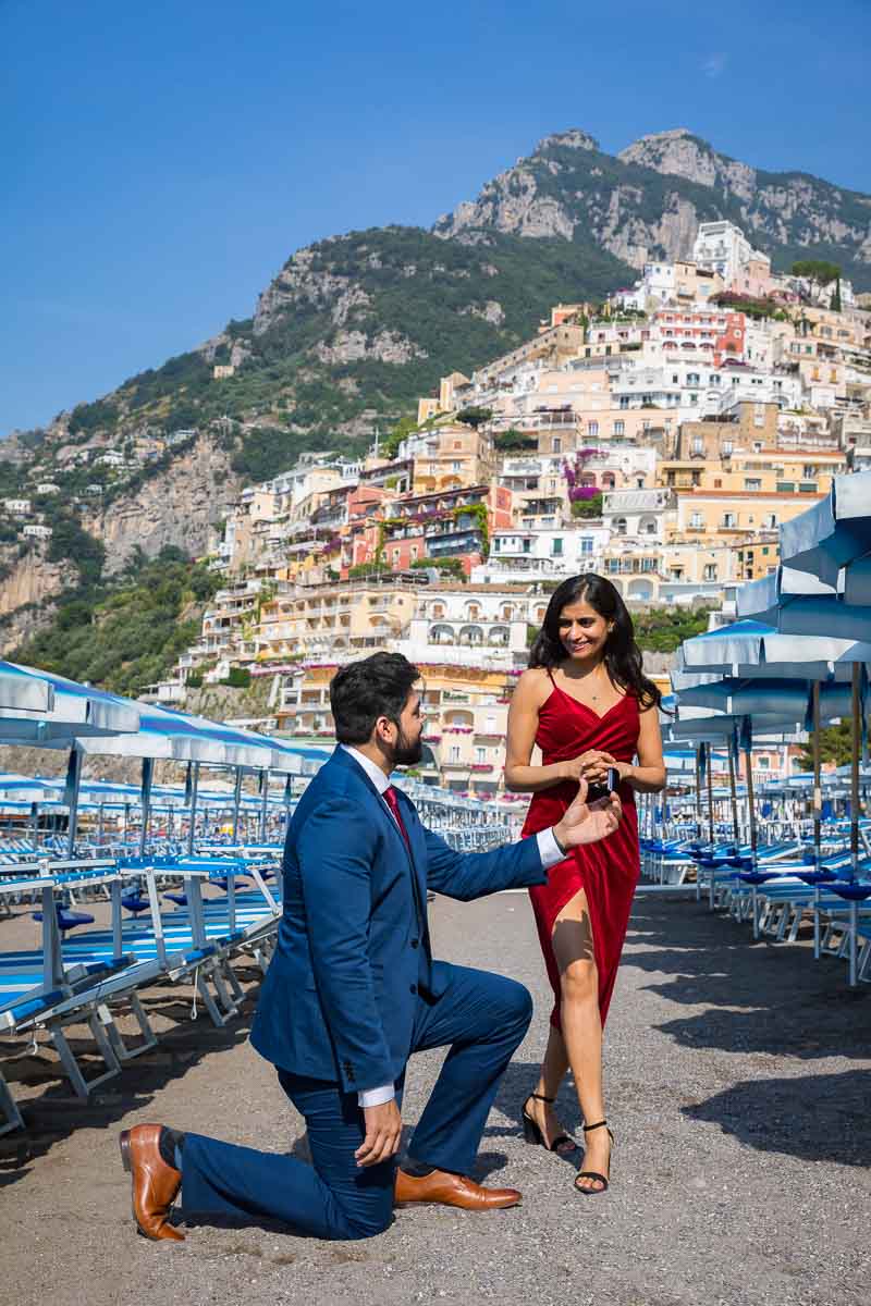 Man proposing on the beach in front of the town of Positano among blue and white beach umbrellas found on the beach. Wedding proposal