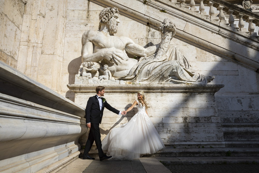 Newlywed couple walking together under a white marble statue in Rome
