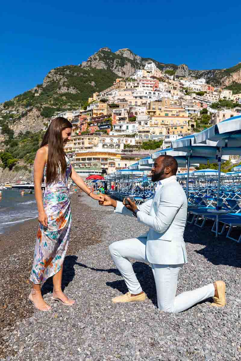 Proposing in Positano. Knee down surprise wedding proposal right on the beach near beach umbrellas and with the vertical town in the background 