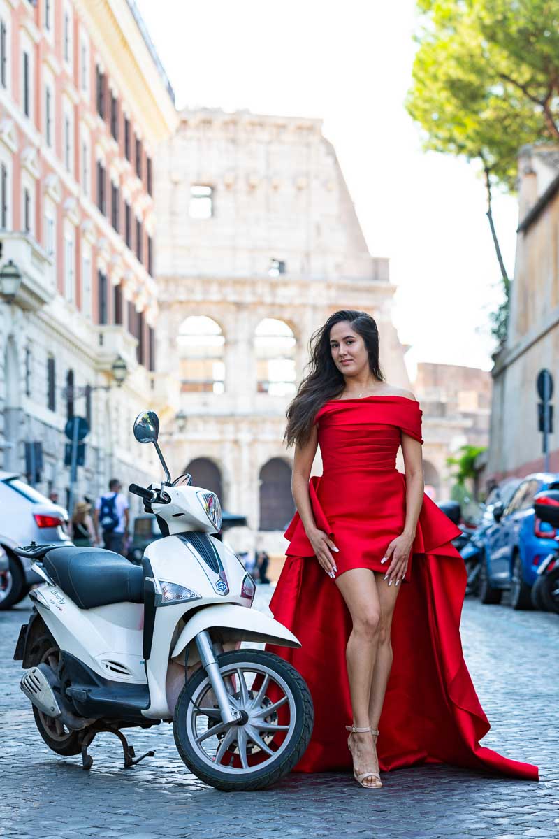 Model wearing a bright red dress posing next to a scooter with the Coliseum in the background 