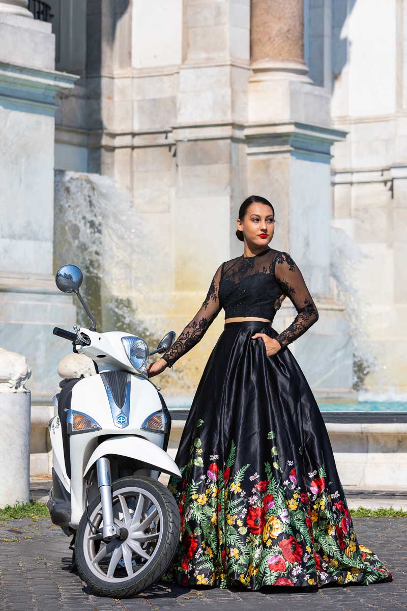 Female model wearing a black dress standing next to a white scooter in front of a scenic water fountain located in Rome's Janiculum hill