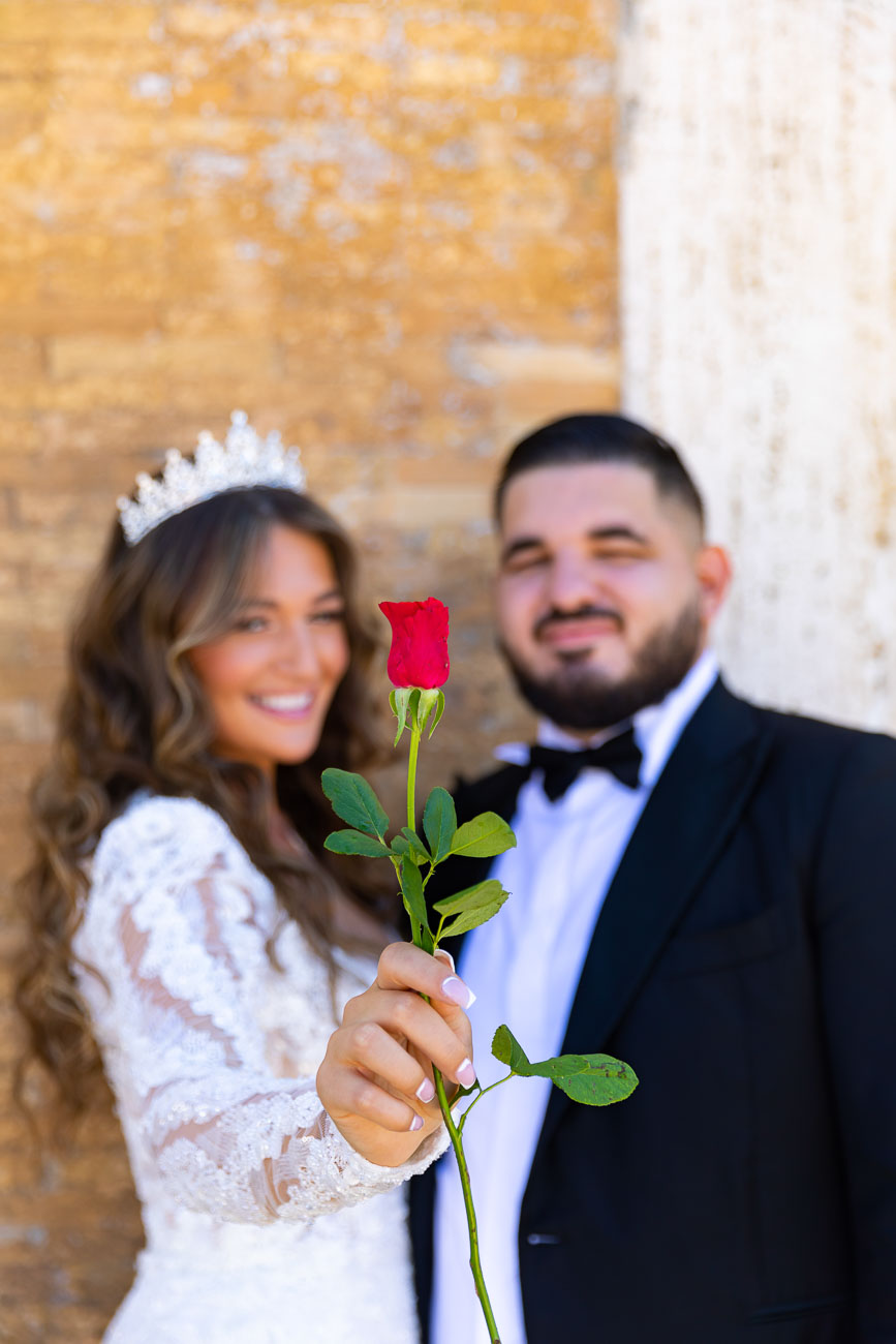 Bride holding a red rose in front for the camera with the bride and groom out of focus in the background