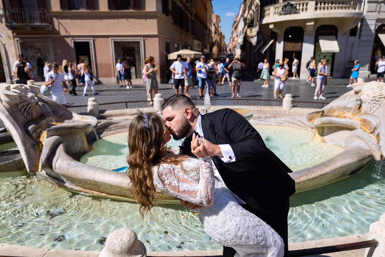 Groom kissing the bride in font of the barcaccia water fountain found at the bottom of the Spanish steps with the background people out of focus