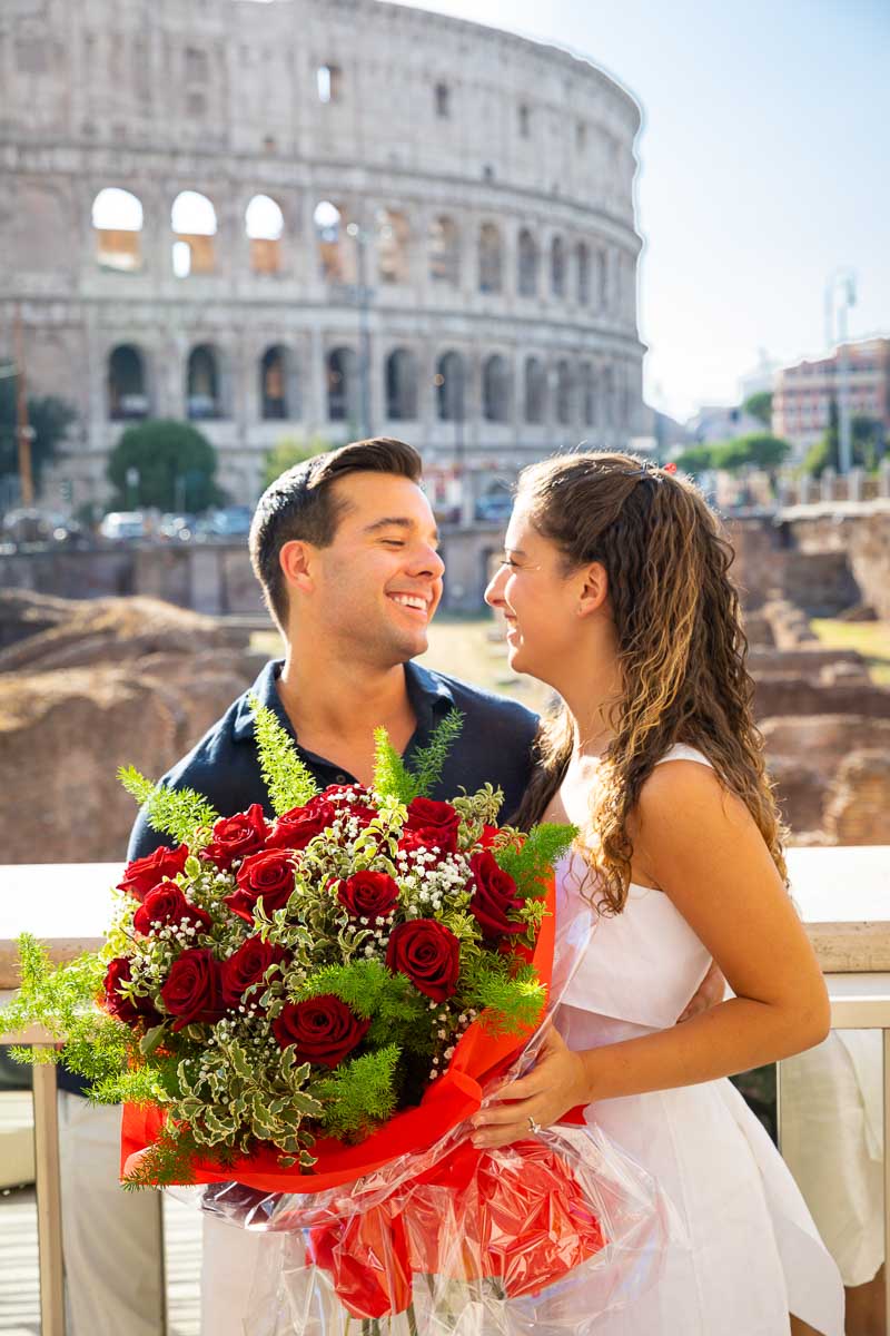 Coupe portrait posing with a large bouquet of red roses in front of the Colosseum taking engagmenet pictures