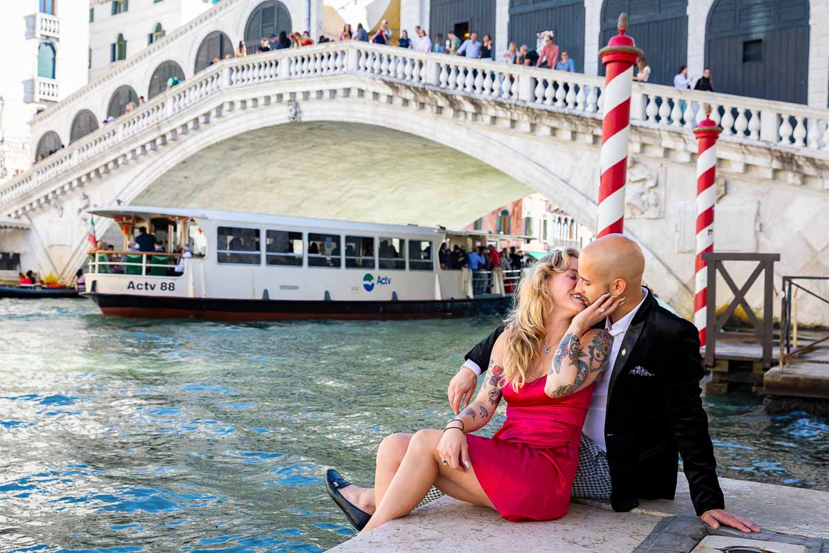 Couple sitting down next to the grand canal in Venice with the Rialto bridge in the background
