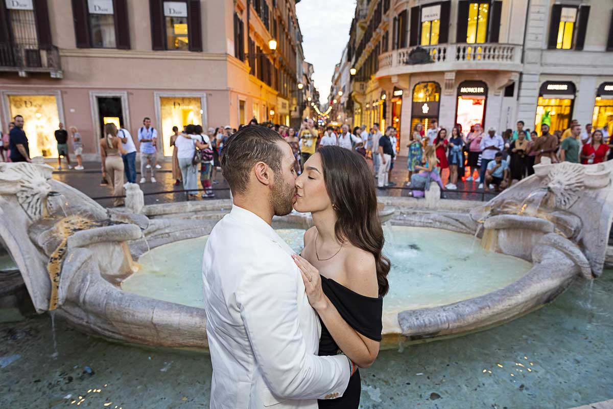 Kissing by the Barcaccia water fountain found at the bottom of the Spanish steps in Rome Italy as the lights start to turn on 
