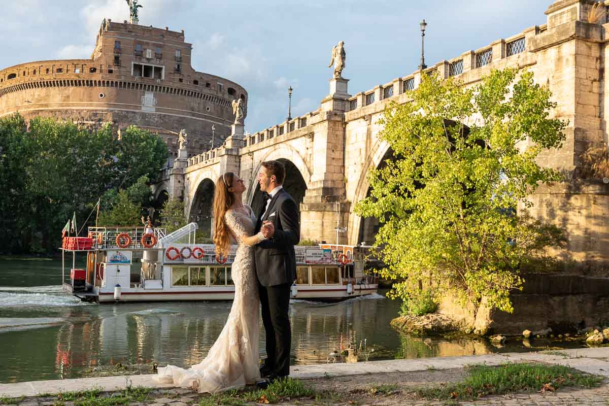 Groom and bride posed together under Castel Sant'Angelo bridge with a view of the Castle while a boat is rolling along on the river in the background