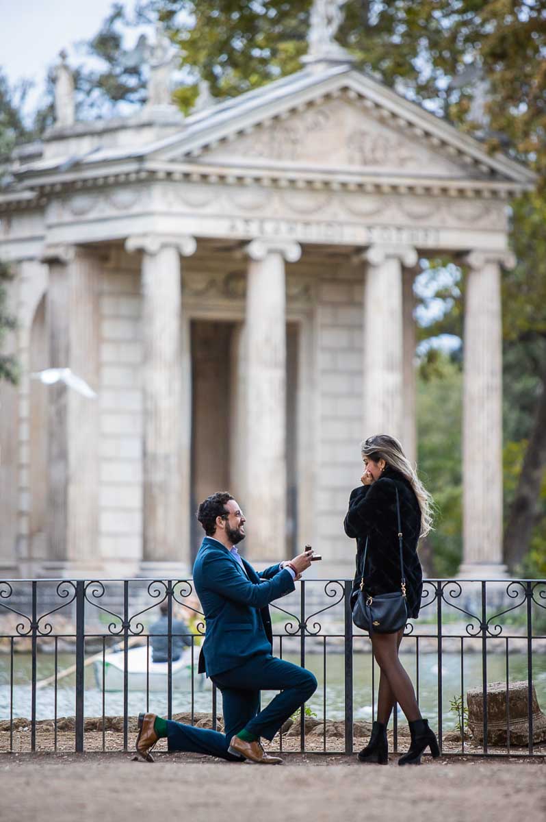 Knee down wedding marriage proposal candidly photographed from a distance in Villa Borghese park. Asclepius Temple proposal