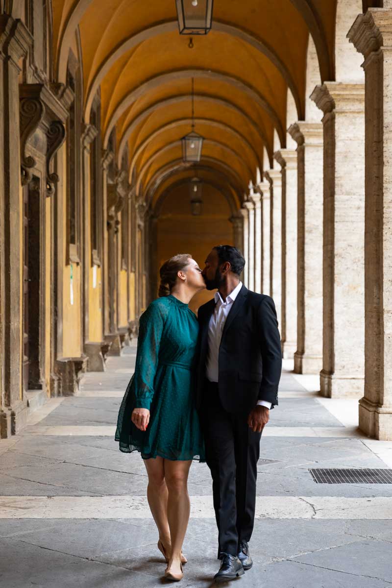 Engagement couple walking while kissing under roman porticos 