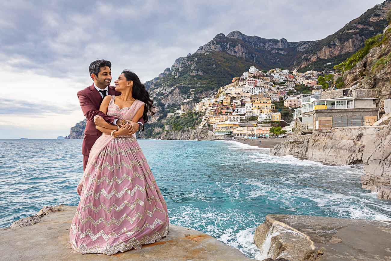 Engagement couple photo taken next to the beach with the vertical town of Positano in the far distance as backdrop 