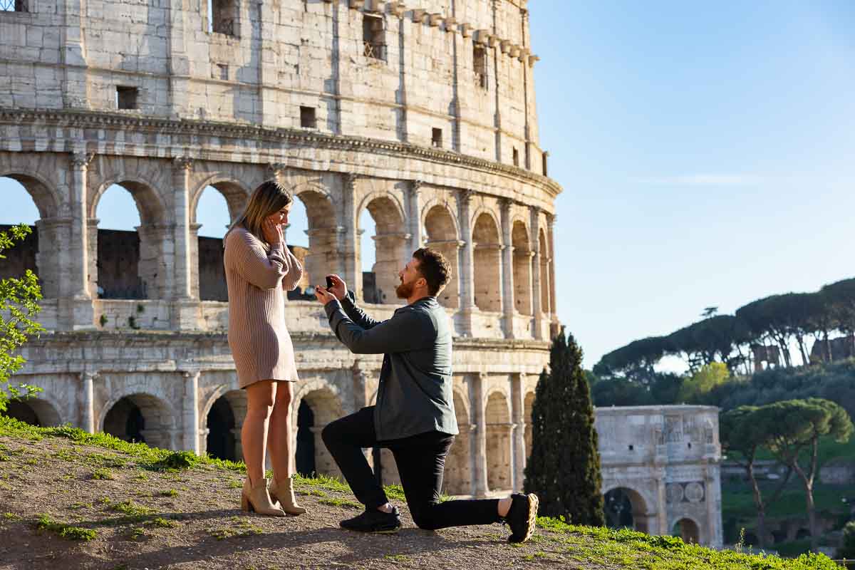 Man knee down wedding marriage proposal photography taken at the roman Colosseum. Surprise engagement photo session in Rome Italy