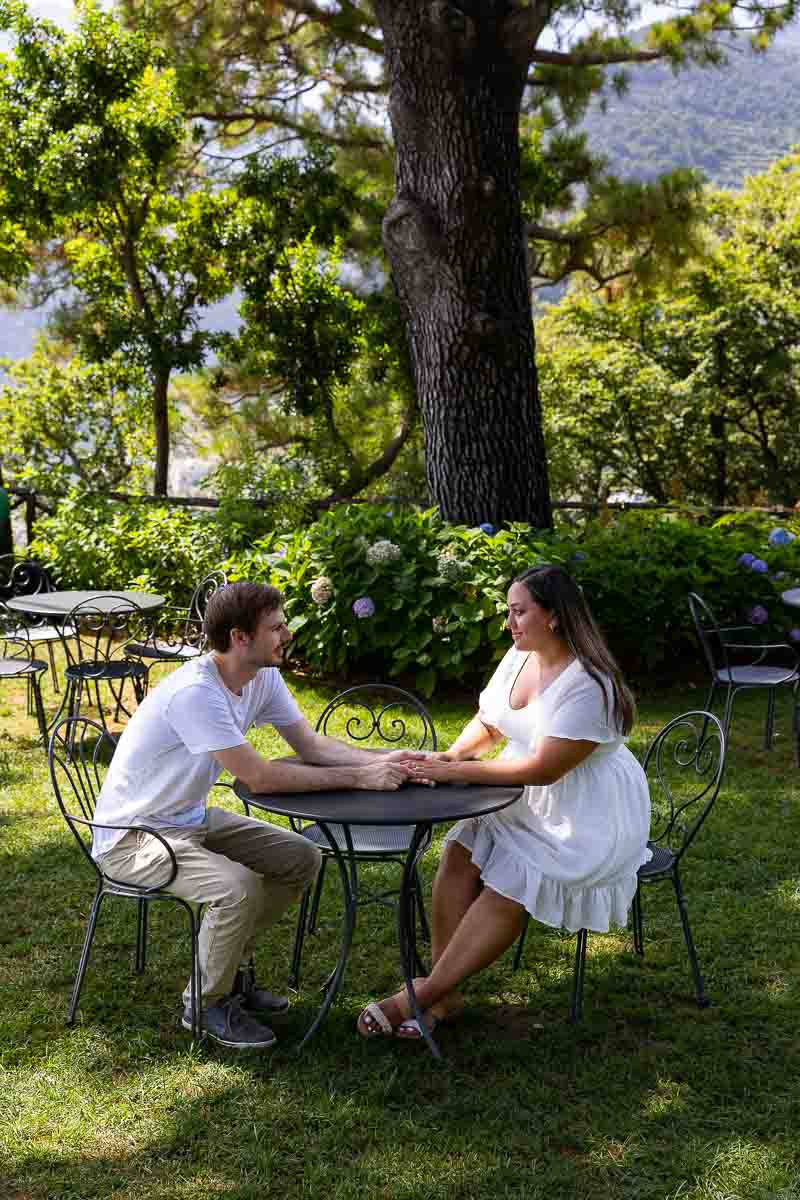 Couple sitting down holding hands in a green park with chairs chairs and trees