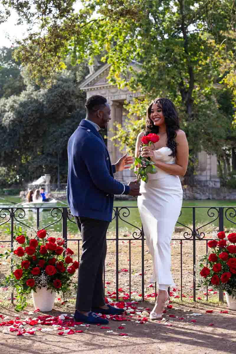 Offering red roses during a surprise proposal in Rome's Villa Borghese lake with an ancient temple in the background 