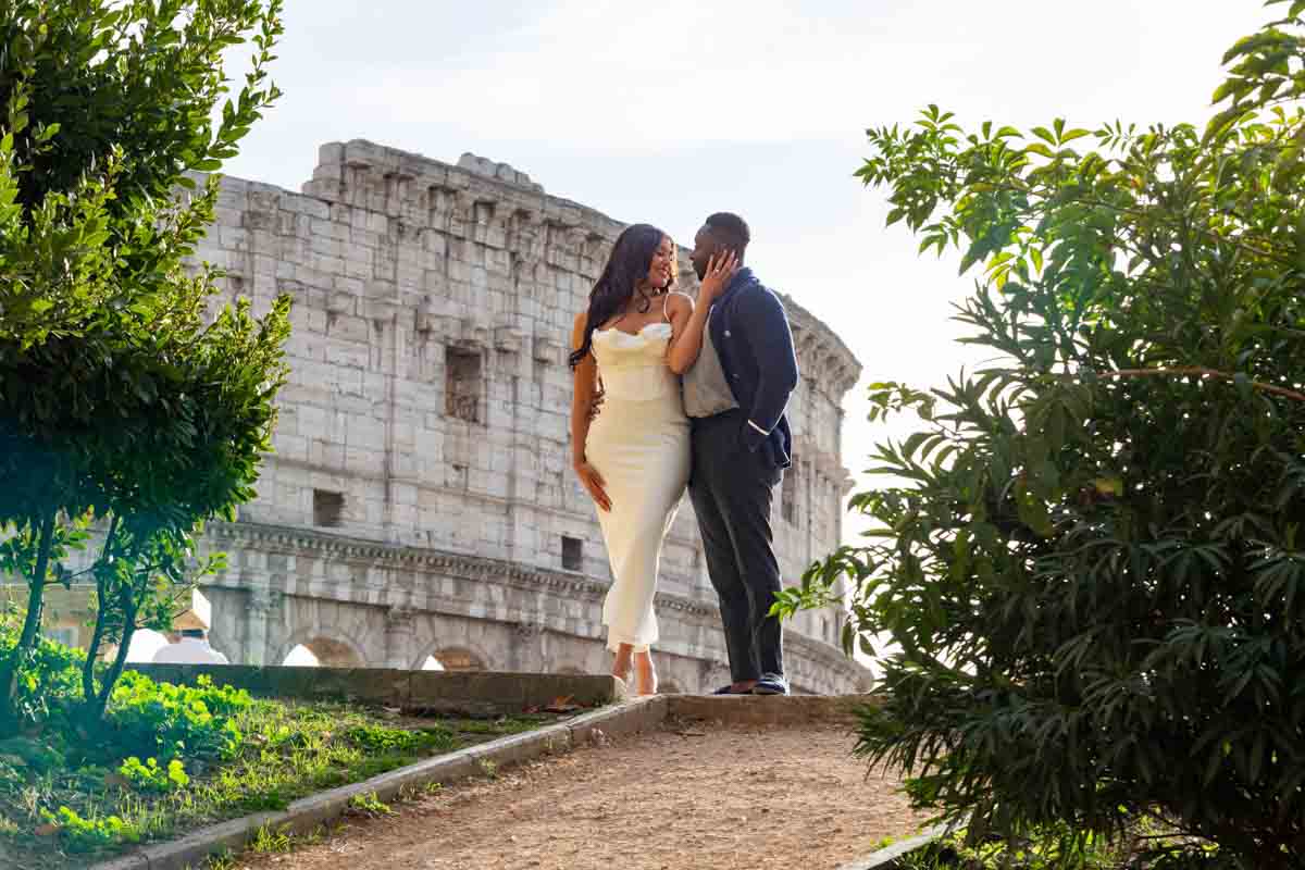 Couple standing before the ancient roman colosseum monument while taking unique and creative photographs of the their engagement in Rome