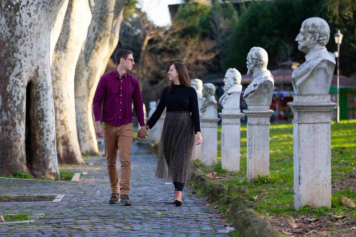 Walking in Rome. Couple holding hands and walking by statue busts and typical roman trees 