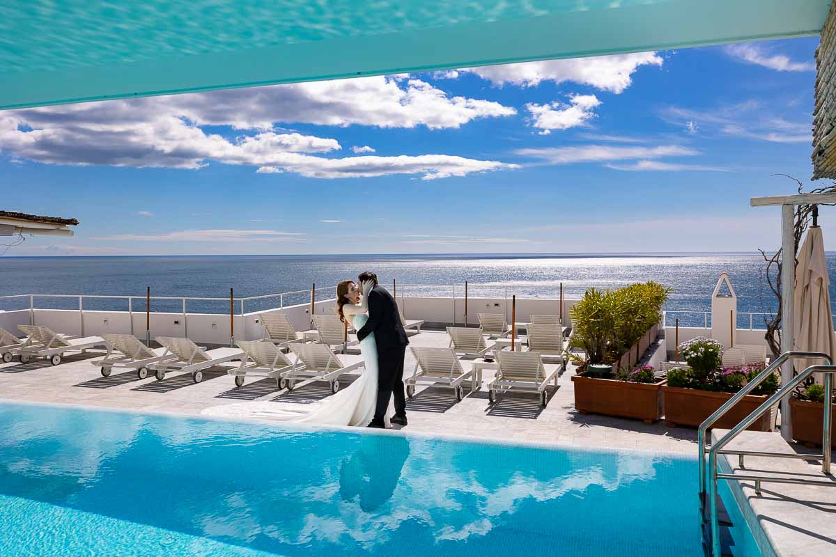 Bride and groom on a destination wedding before the amazing and incredible view of the Amalfi coast with white fluffy clouds and blue sky. Not to mention an amazing infinity swimming pool