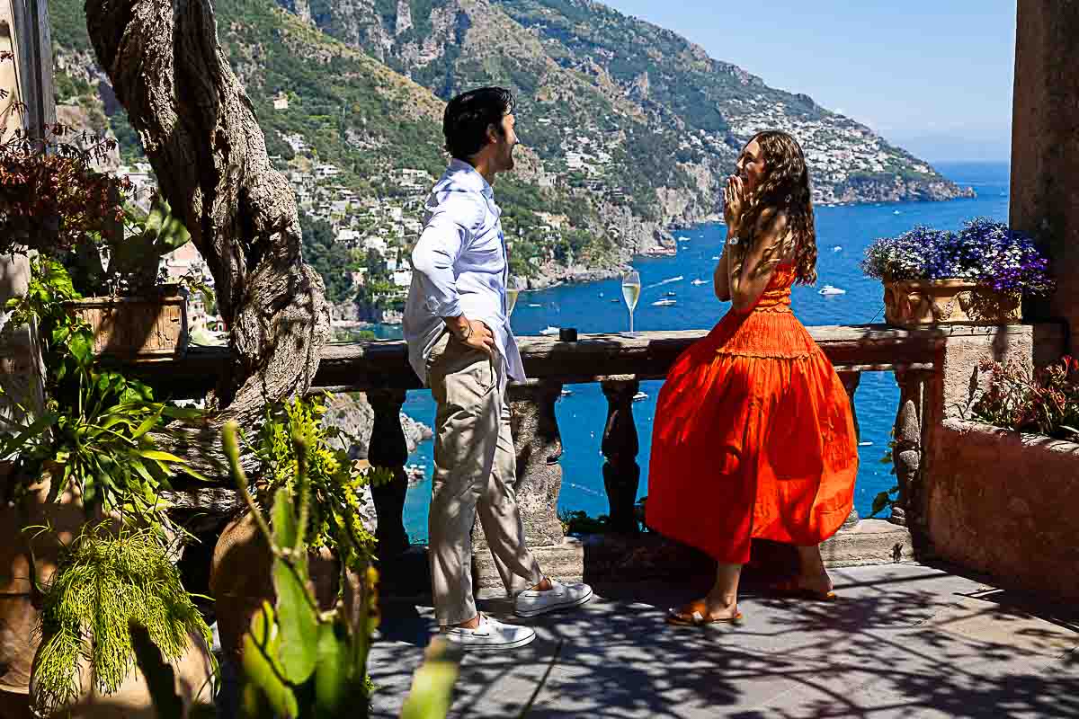 Joyful moment on a sunlit terrace after a surprise engagement, with coastal cliffs and sea views in the distance
