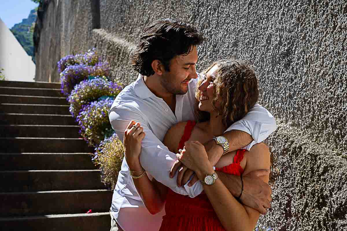 Couple smiling warmly at each other while embracing on a quaint stone staircase decorated with colorful flowers