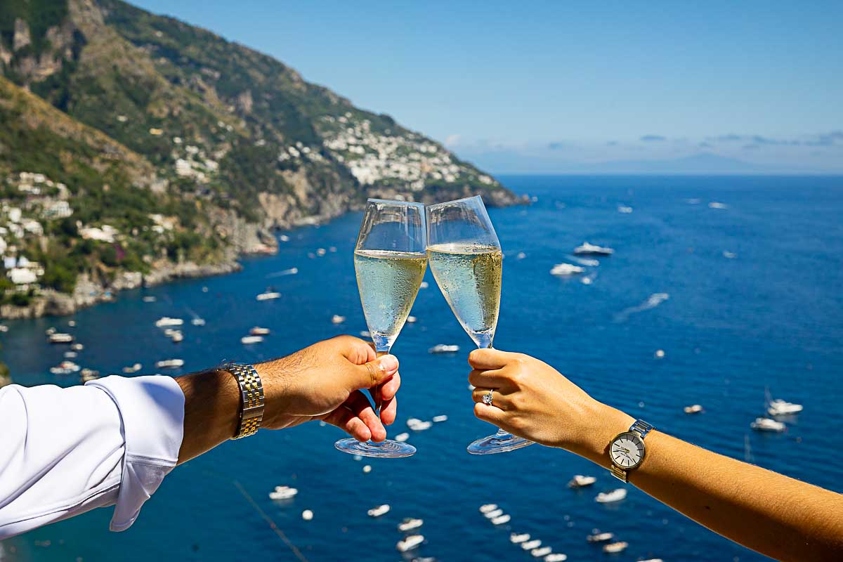 Two glasses tilting in a celebratory toast for the newly engaged with sparkling prosecco, overlooking the shimmering sea at sunset. Image take during a professional Positano Proposal Photographer session