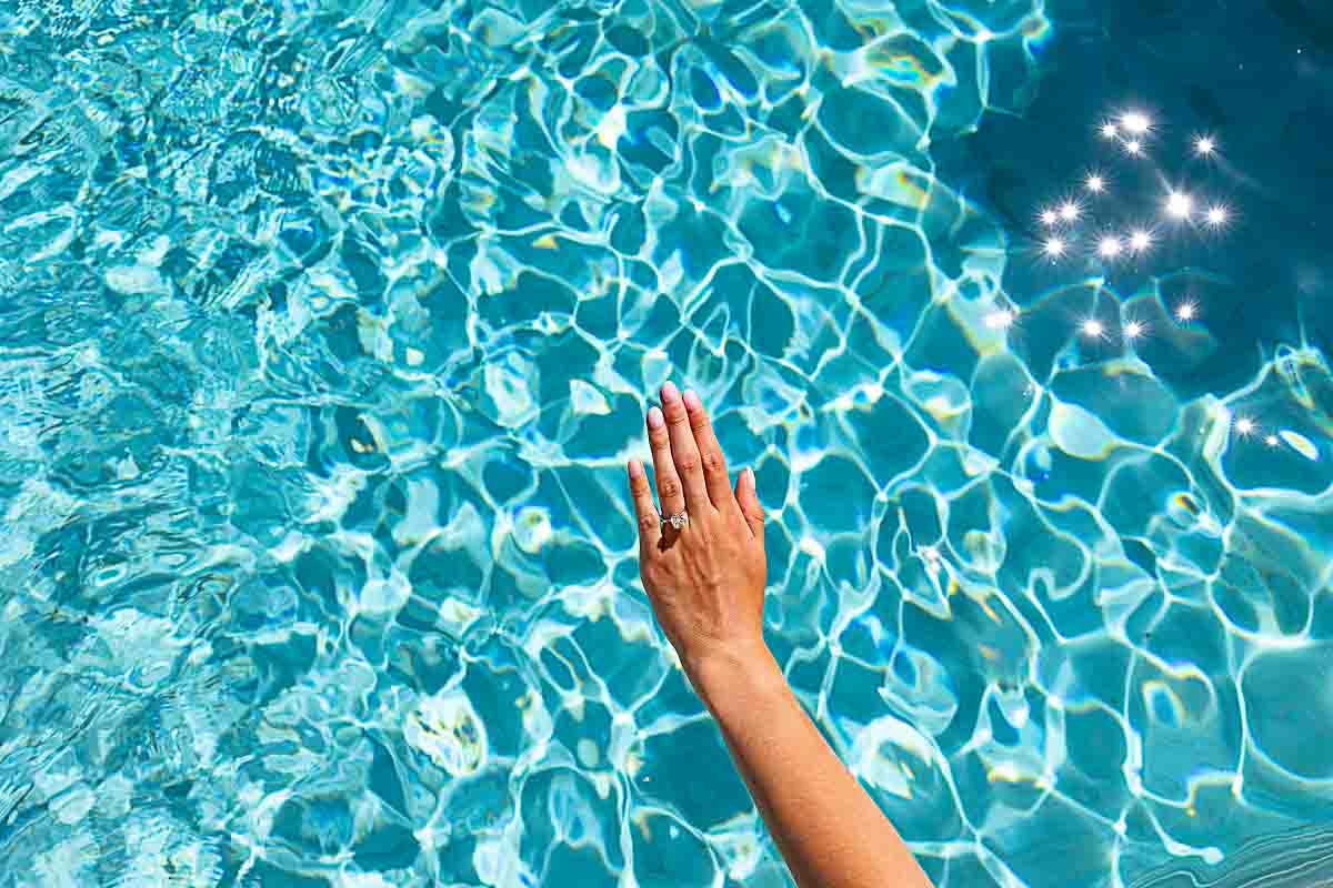 Close-up of an engagement ring held just above a vibrant turquoise swimming pool, the colorful water creating a striking backdrop
