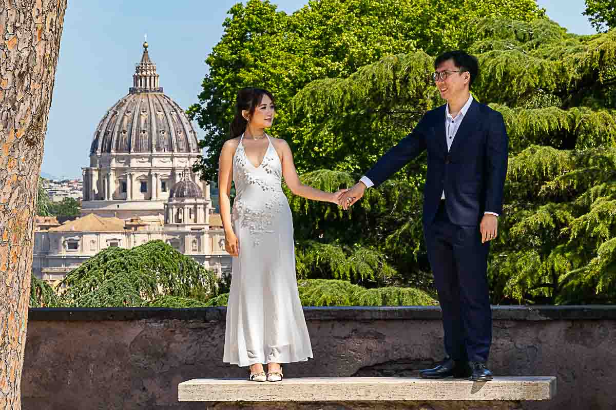 Holding hands while standing on a marble bench in front of the Vatican church basilica dome in the distance 