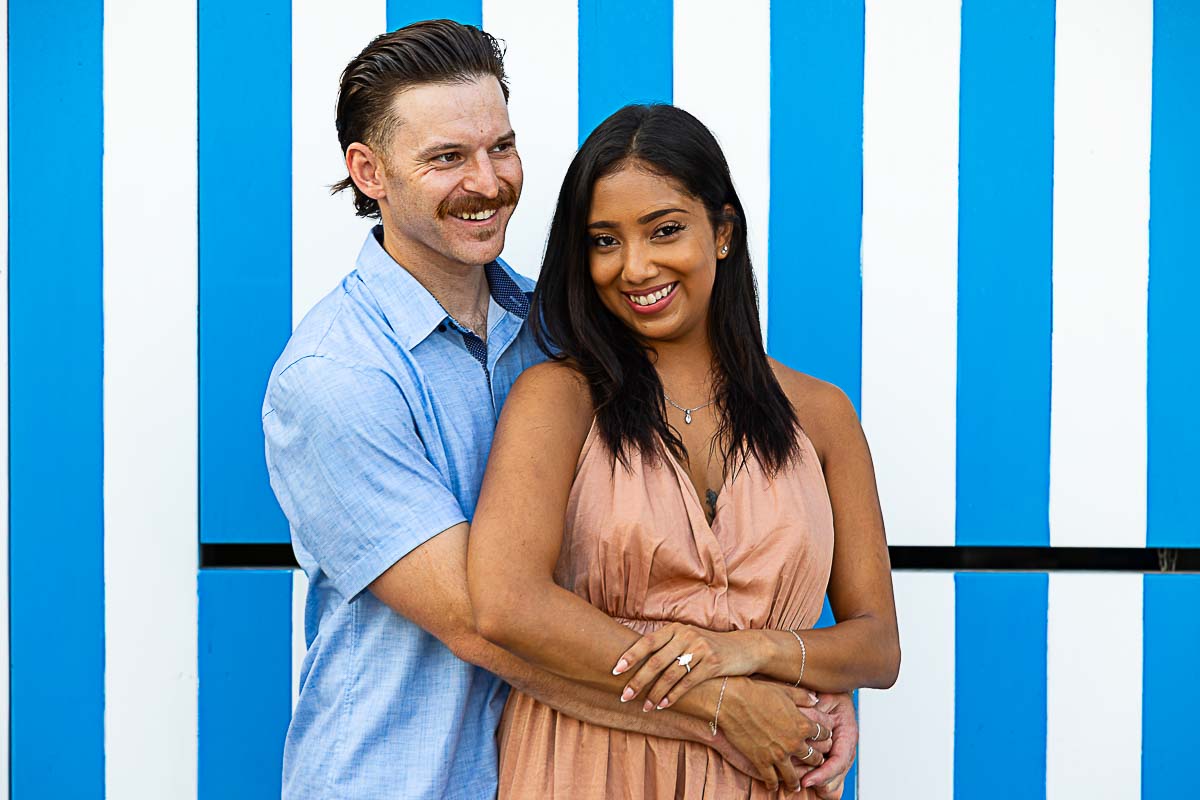 Close up portrait photo of engaged couple in front of blue and white typical beach cabin