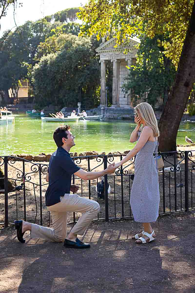 Man proposing marriage knee down in front of the Temple of Aesculapius and the lake candidly photographed from a distance by a professional photographer 