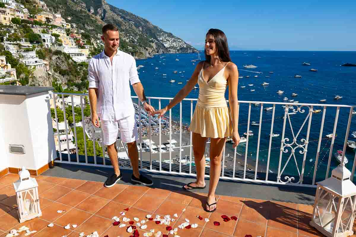 Couple holding hand in hand while standing in between white lanterns and standing over rose petals on the ground with the sea in the background 