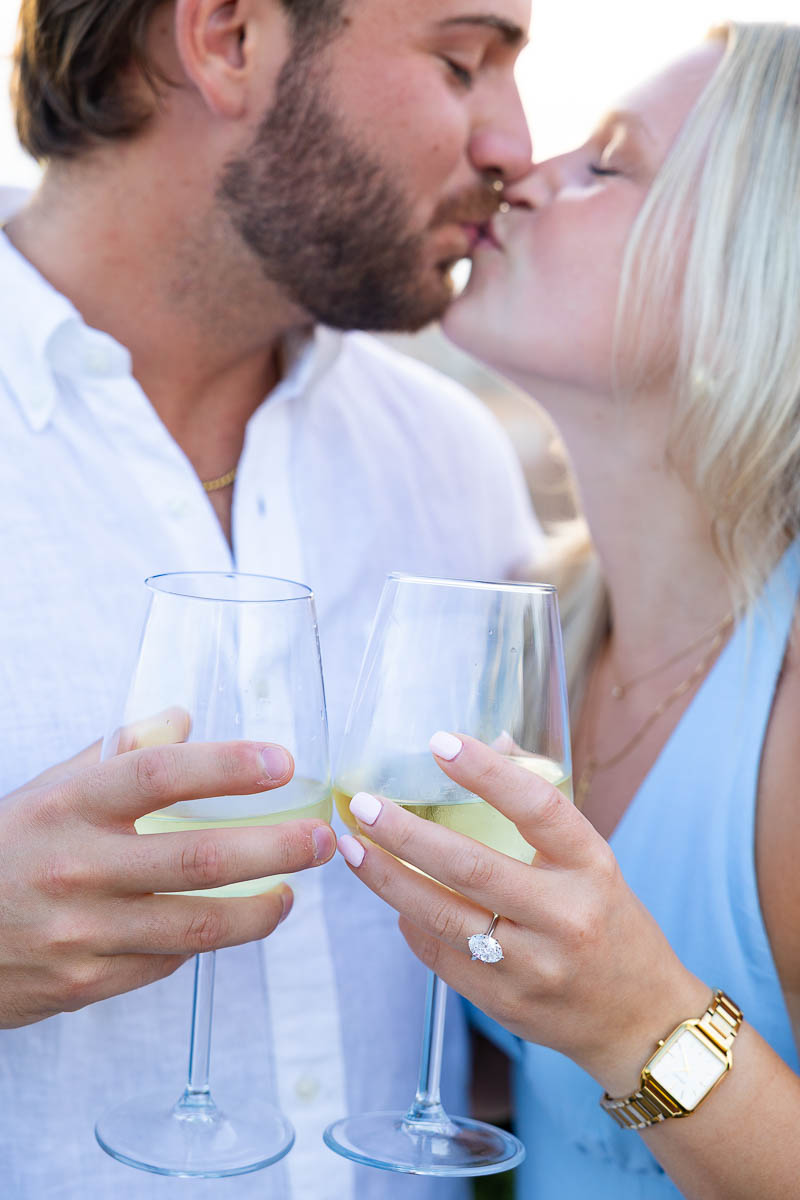 Close-up of the wine glasses showing the engagement ring with the subject kissing in the background 